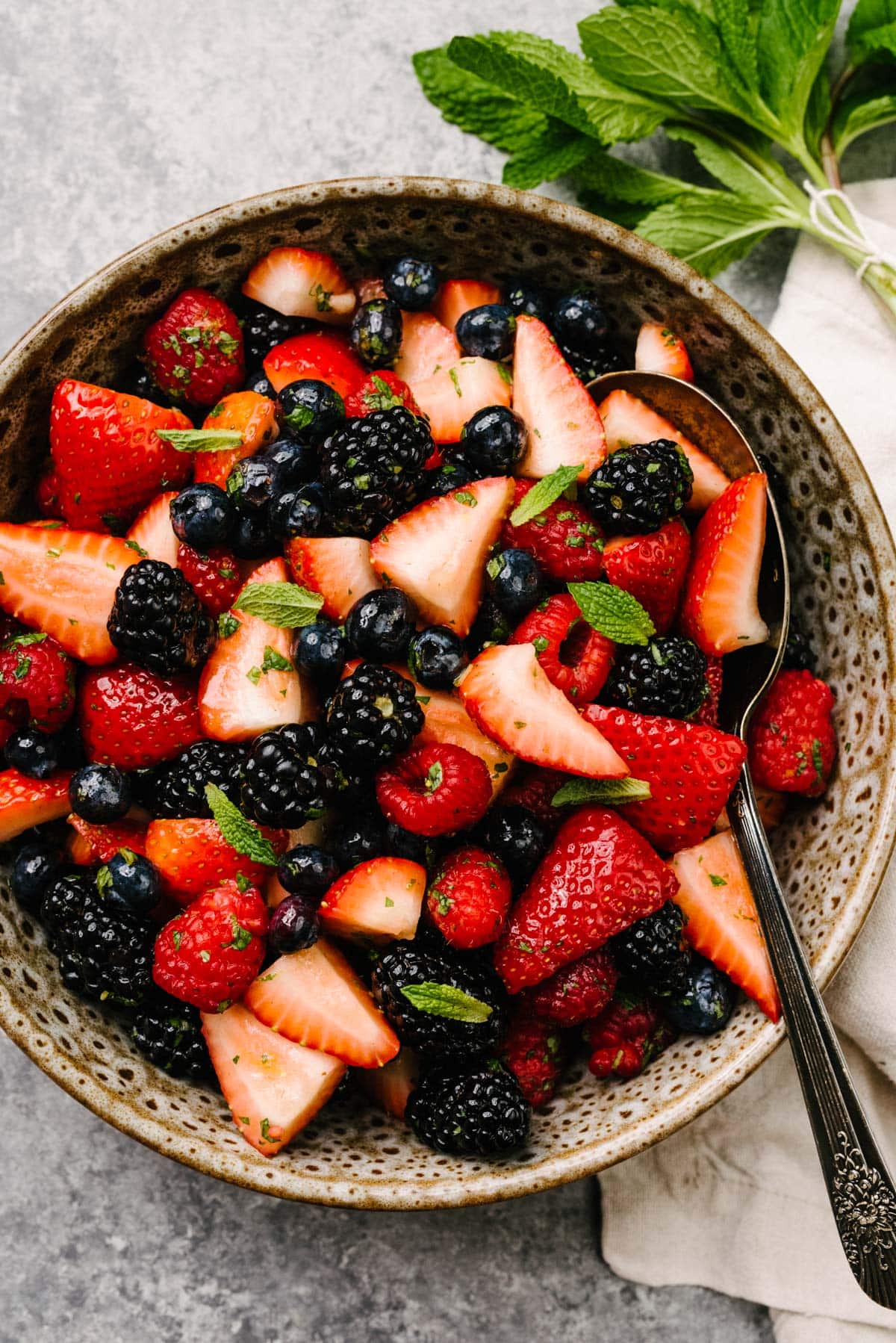 Berry fruit salad with strawberries, blueberries, blackberries, and raspberries tossed with honey mint dressing in a brown speckled bowl on a concrete background; cream linen napkin and bundle of fresh thyme to the side.