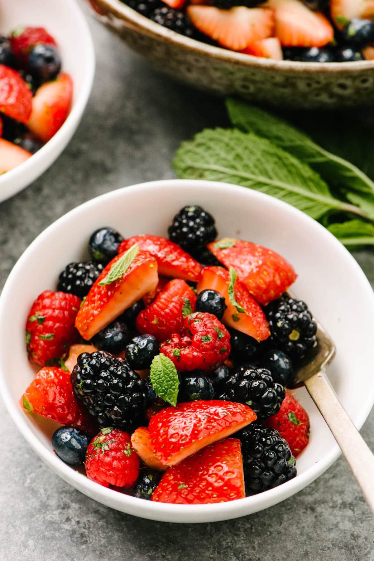 Side view, a fork tucked in a white bowl filled with a fresh berry salad made with strawberries, blackberries, blueberries, raspberries, mint, and a honey lemon dressing. A bundle of fresh mint as well as another small and large bowl of salad are in the background.