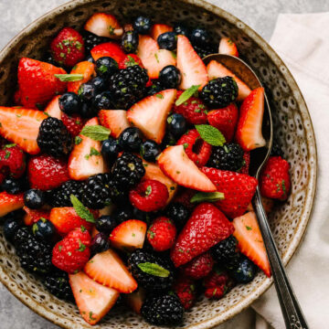 A berry fruit salad made with strawberries, blueberries, blackberries, raspberries, mint, and lemon honey dressing in a brown speckled bowl with a cream linen napkin to the side.