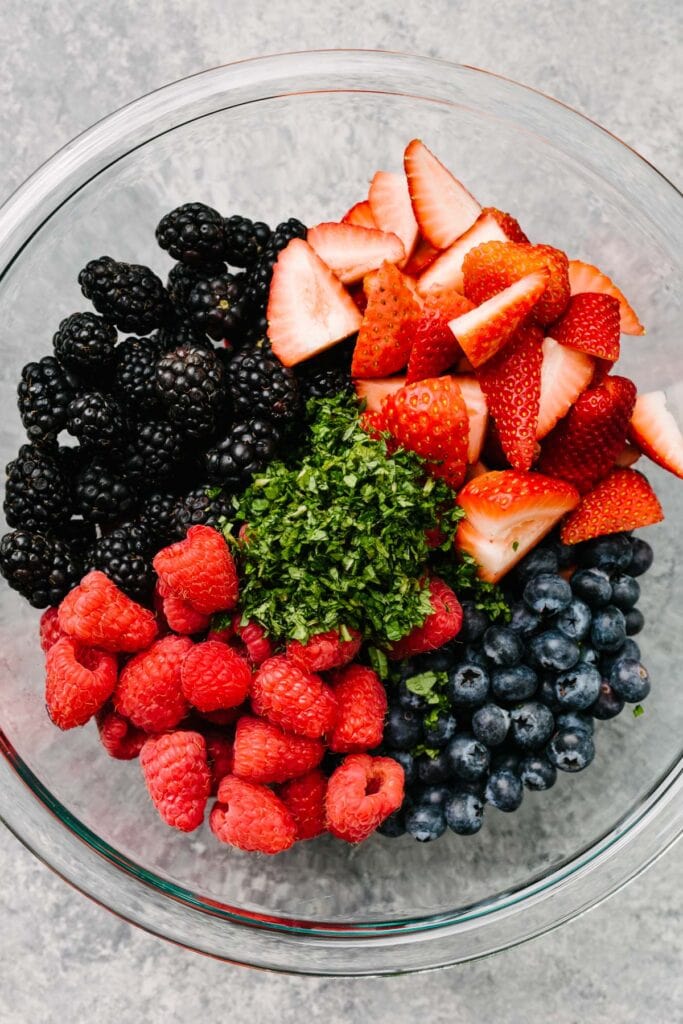 Chopped strawberries, blueberries, blackberries, and raspberries in a large glass mixing bowl with chopped fresh mint.