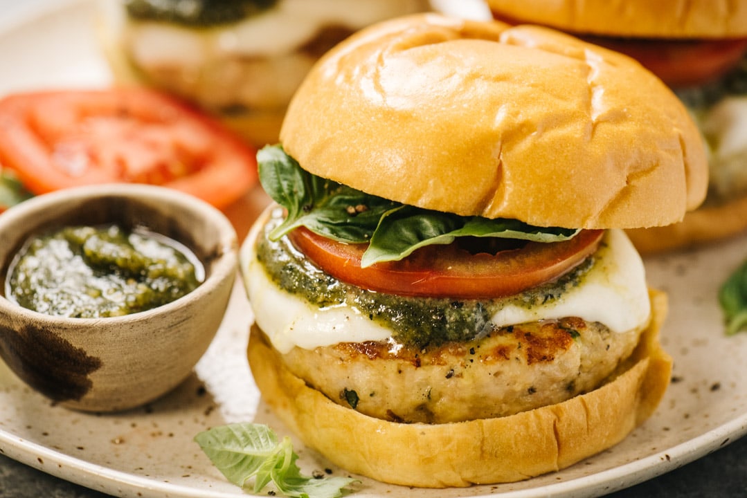Side view, a caprese chicken burger on a tan speckled plate made with a ground chicken burger topped with fresh mozzarella, pesto sauce, tomato slices, and fresh basil on a brioche bun. A small bowl of pesto and additional burgers are in the background.
