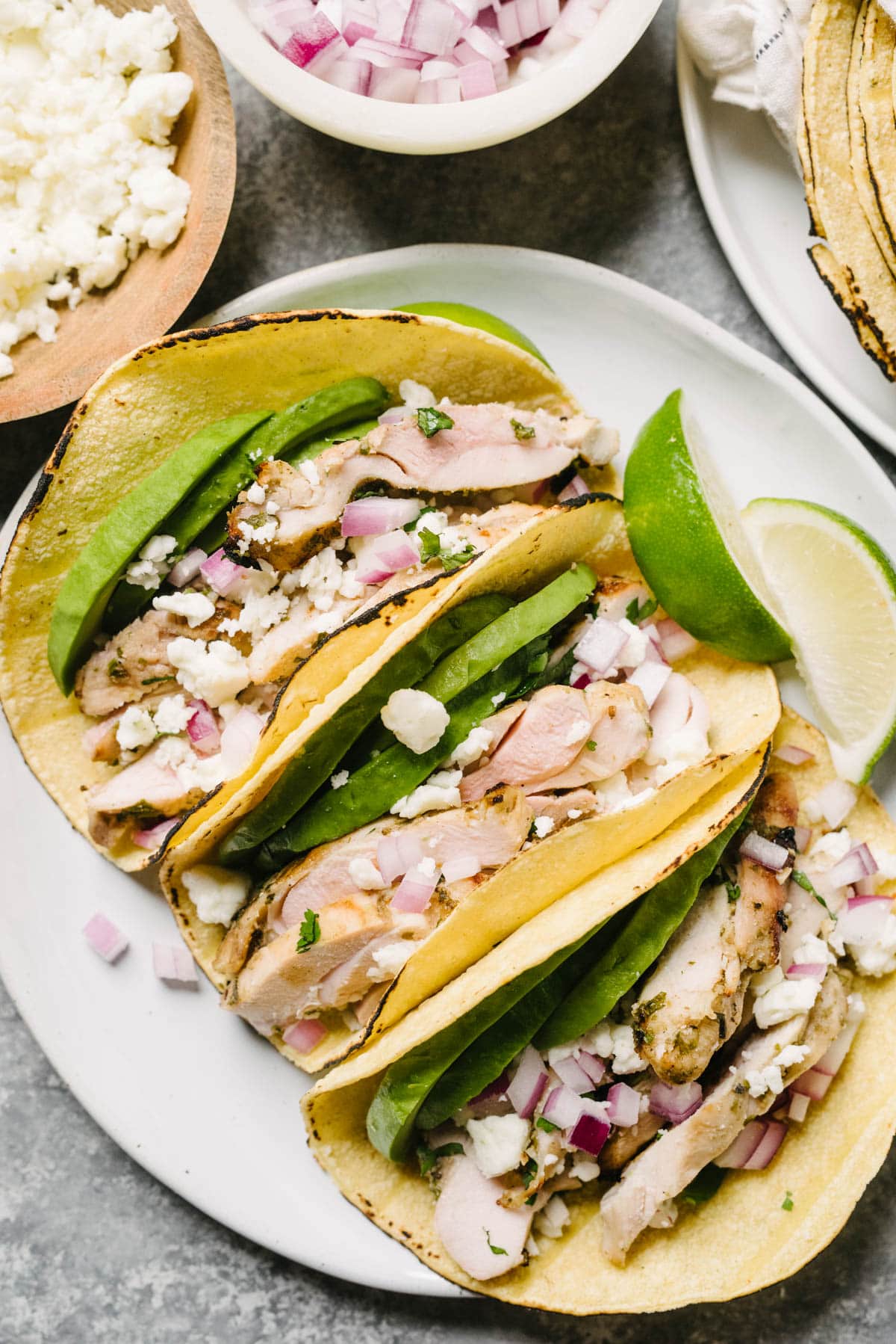 Three cilantro lime chicken tacos on a plate with garnishes; the plate is surrounded by bowls of red onions, cojita cheese, and corn tortillas.