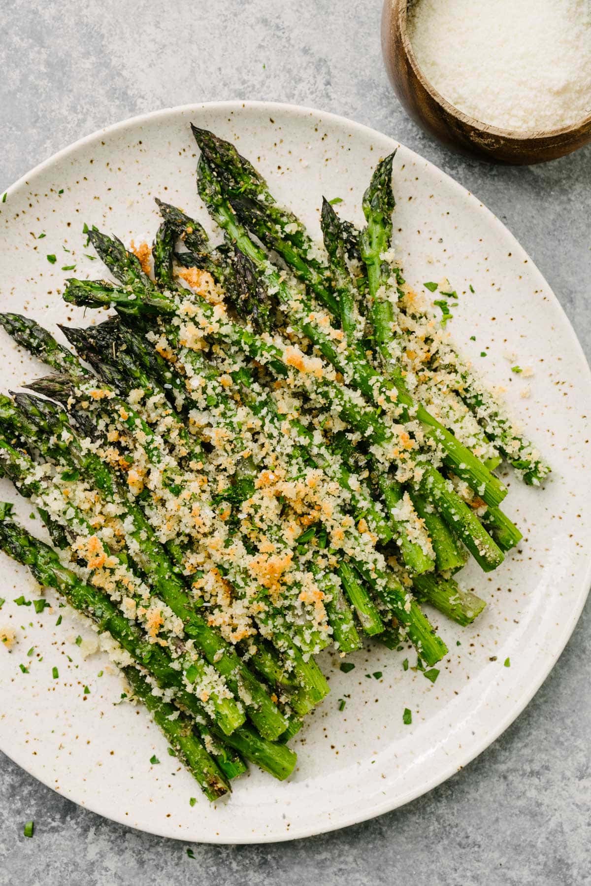Roasted asparagus with parmesan on a brown speckled plate on a concrete background, with a small bowl of grated parmesan cheese to the side.