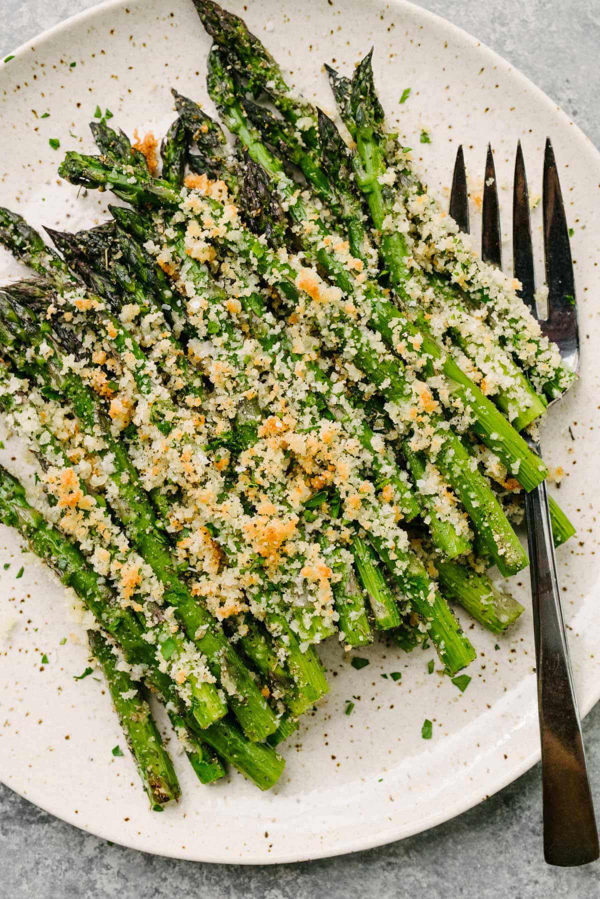 A serving fork tucked under a stack of roasted asparagus topped with a crispy parmesan and breadcrumb topping on a tan speckled plate.