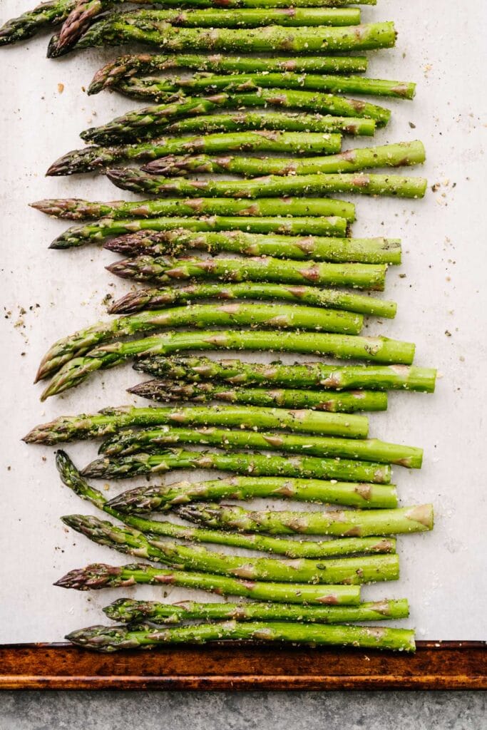 Asparagus spears tossed with olive oil, garlic powder, Italian seasoning, salt, and pepper arranged in a single layer on a parchment lined baking sheet.