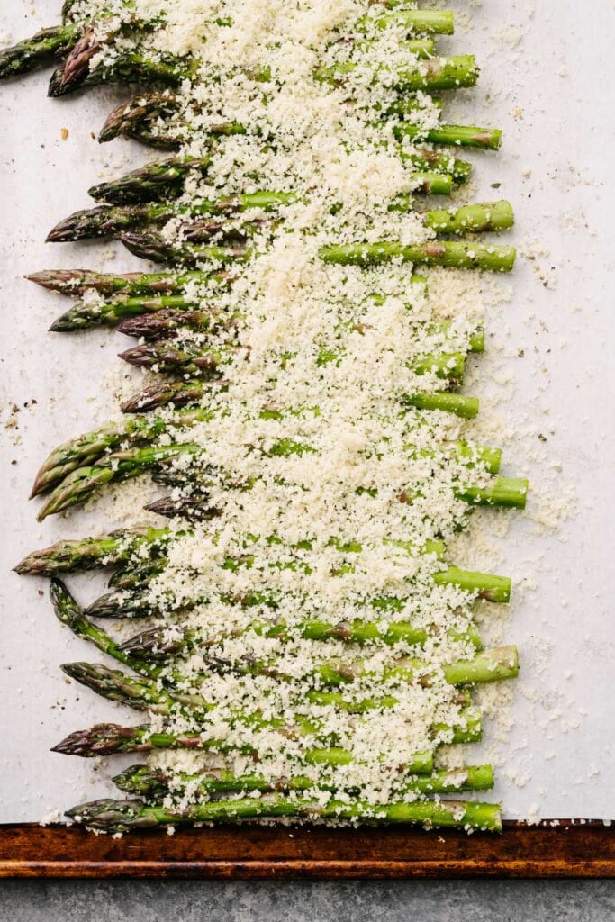 Asparagus spears topped with a parmesan cheese and bread crumb topping on a parchment lined baking sheet, before going into the oven.