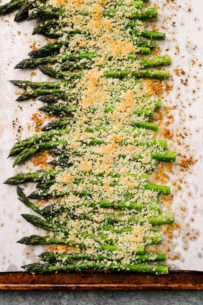 Roasted asparagus spears topped with a crispy garlic, parmesan, and breadcrumb topping on a parchment lined baking sheet.