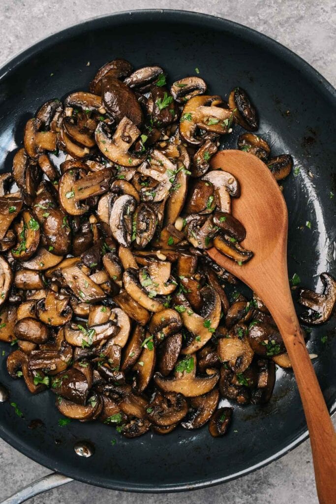 A wood spoon tucked into a skillet of golden brown sautéed mushrooms, garnished with fresh chopped parsley.