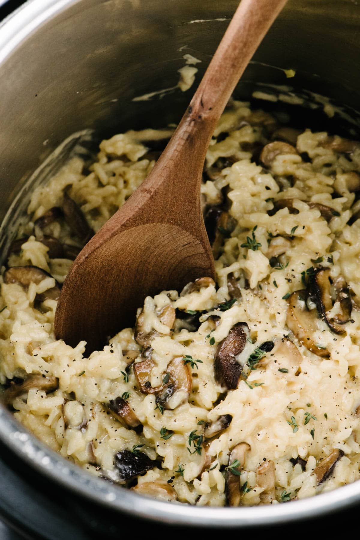 Side view, a wood spoon tucked into mushroom risotto in an Instant Pot, garnished with fresh thyme leaves.