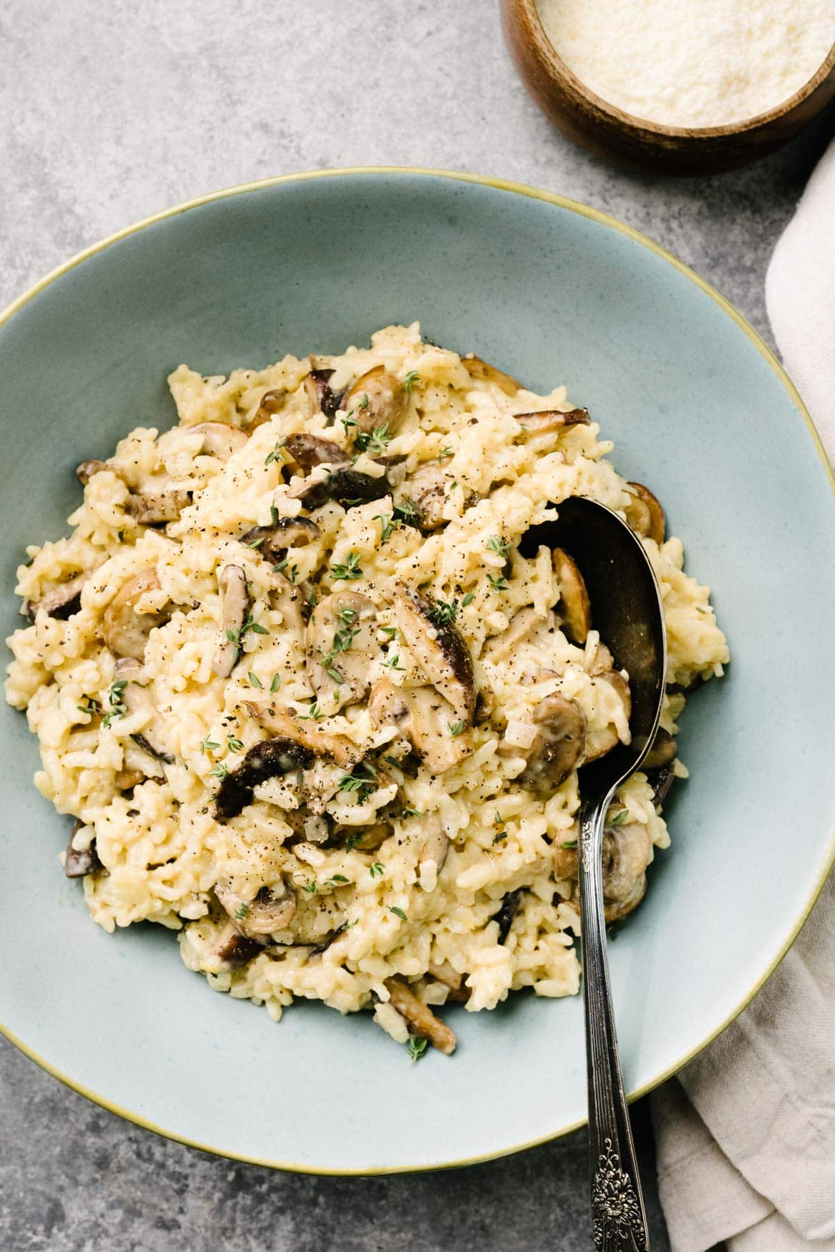 Instant Pot mushroom risotto in a blue serving bowl, garnished with fresh thyme. A serving spoon is tucked into the risotto, with a cream linen napkin and small wood bowl of grated parmesan cheese to the side.