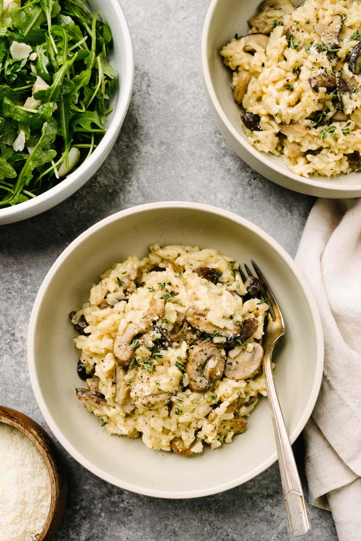 Two bowls of Instant Pot mushroom risotto on a concrete background. An arugula salad, small bowl of grated parmesan cheese, and cream linen napkin surround the bowls.