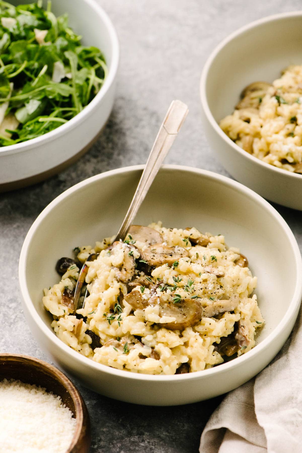 Side view, two bowls of Instant Pot mushroom risotto on a concrete background, surrounded by an arugula salad, cream linen napkin, and small wood bowl of grated parmesan cheese.