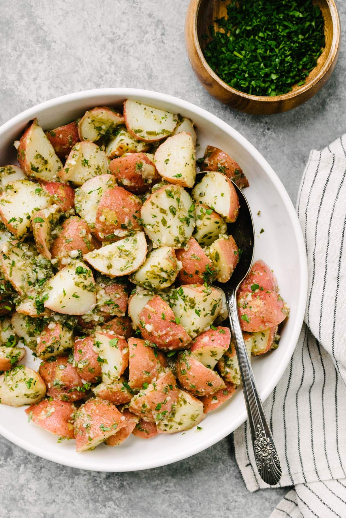 Italian potato salad with vinaigrette dressing in a large white serving bowl with a silver spoon tucked into the potatoes; a striped linen napkin and small bowl of fresh minced herbs to the side.