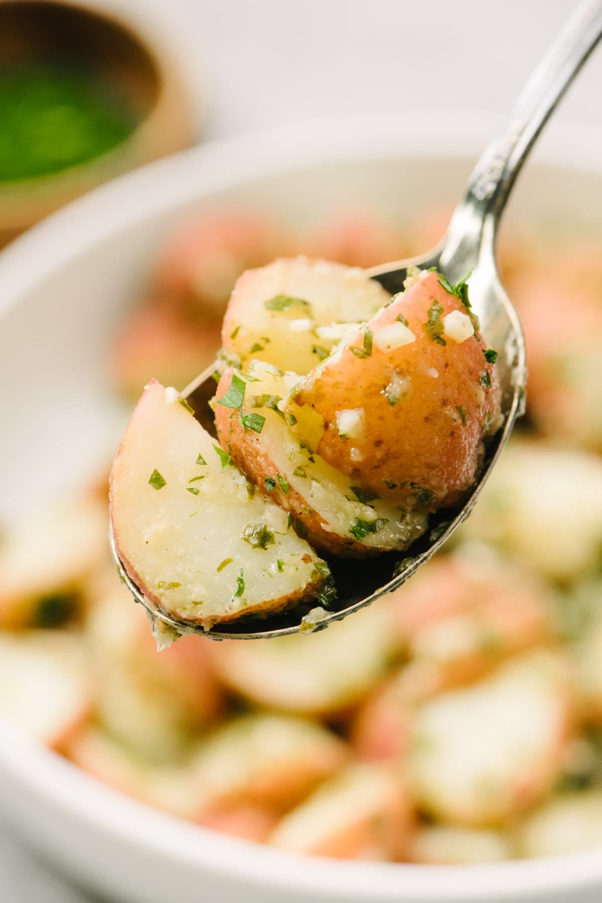 Side view, a serving spoon of no-mayo potato salad hovering over a serving bowl with a small bowl of fresh herbs in the background.
