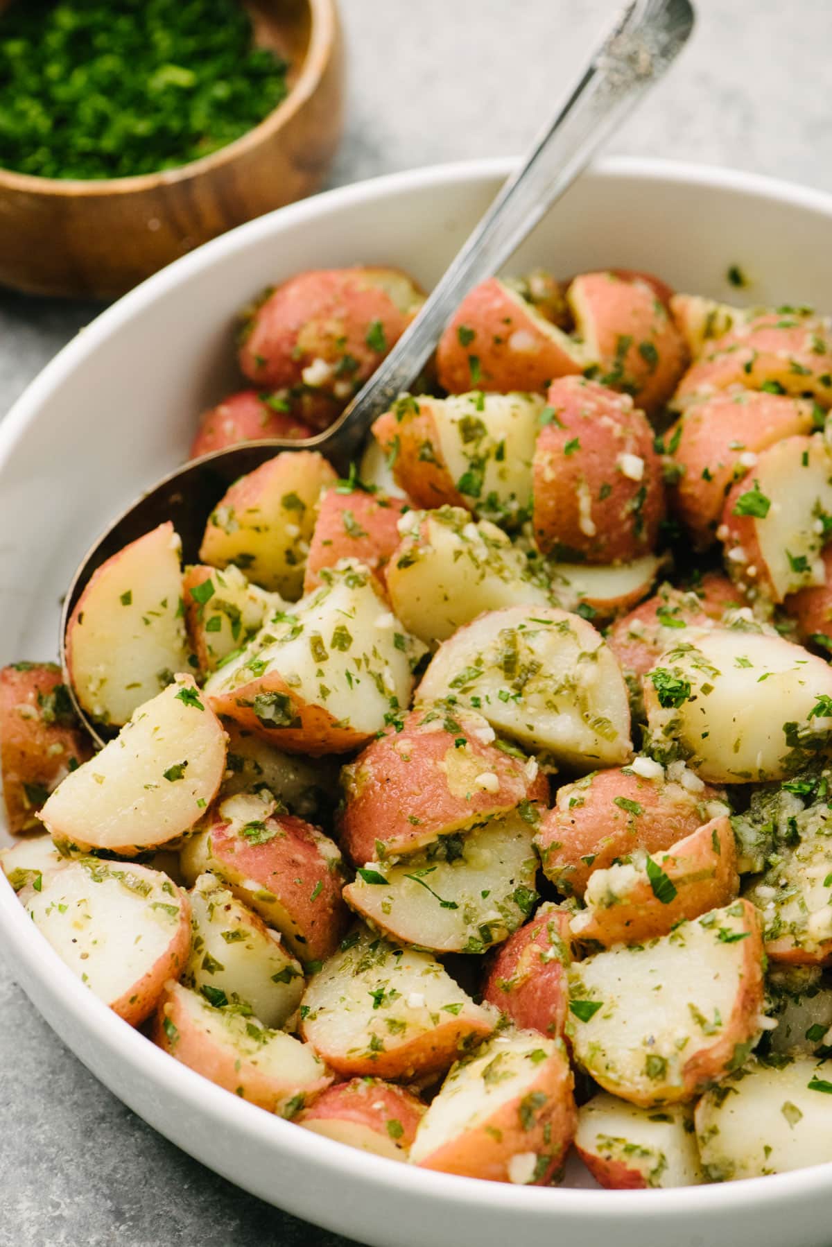 Side view, Italian potato salad with vinaigrette in a large white serving bowl with a small wood bowl of fresh minced herbs in the background.