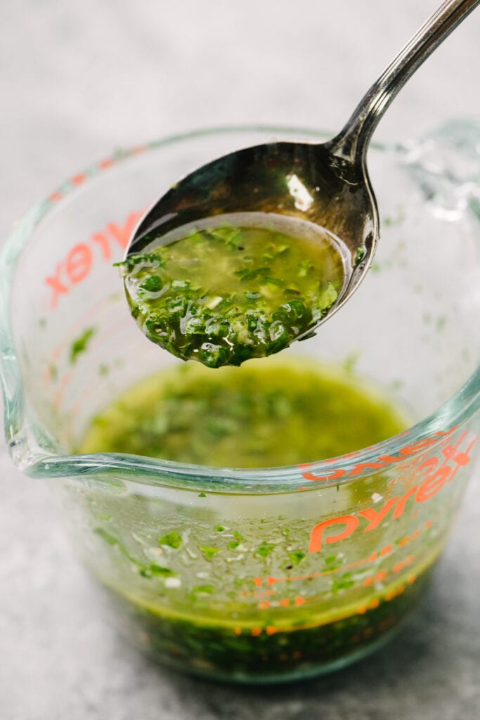Side view, herb vinaigrette running off a spoon and into a 1-cup glass measuring cup.