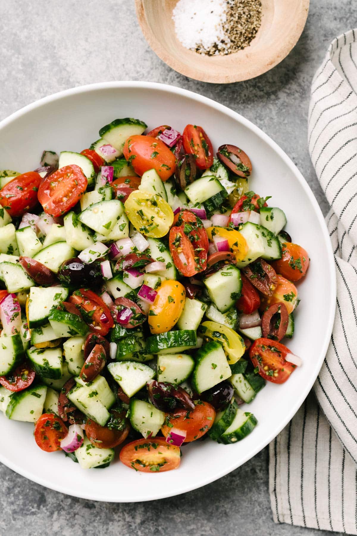 Mediterranean cucumber salad in a large white serving bowl on a concrete background with a striped linen napkin and small wood bowl filled with salt and pepper for seasoning.