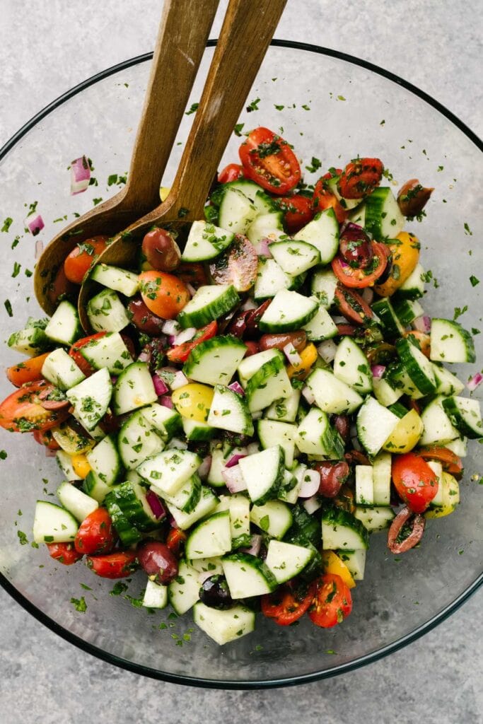 Freshly tossed mediterranean cucumber salad in a large glass mixing bowl with wood serving utensils.