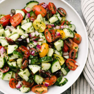 Mediterranean cucumber salad in a large white serving bowl on a concrete background with a striped linen napkin to the side.