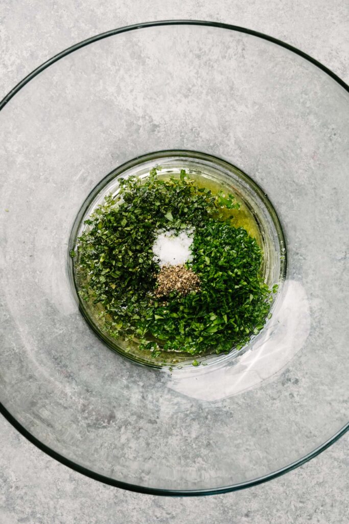 Olive oil, white wine vinegar, fresh parsley, fresh mint, salt, and pepper in a large glass mixing bowl.