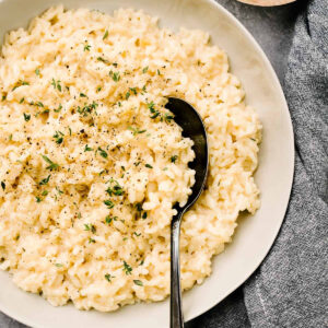 A serving spoon tucked into a bowl of creamy risotto with a bowl of parmesan cheese and dark grey linen napkin to the side.