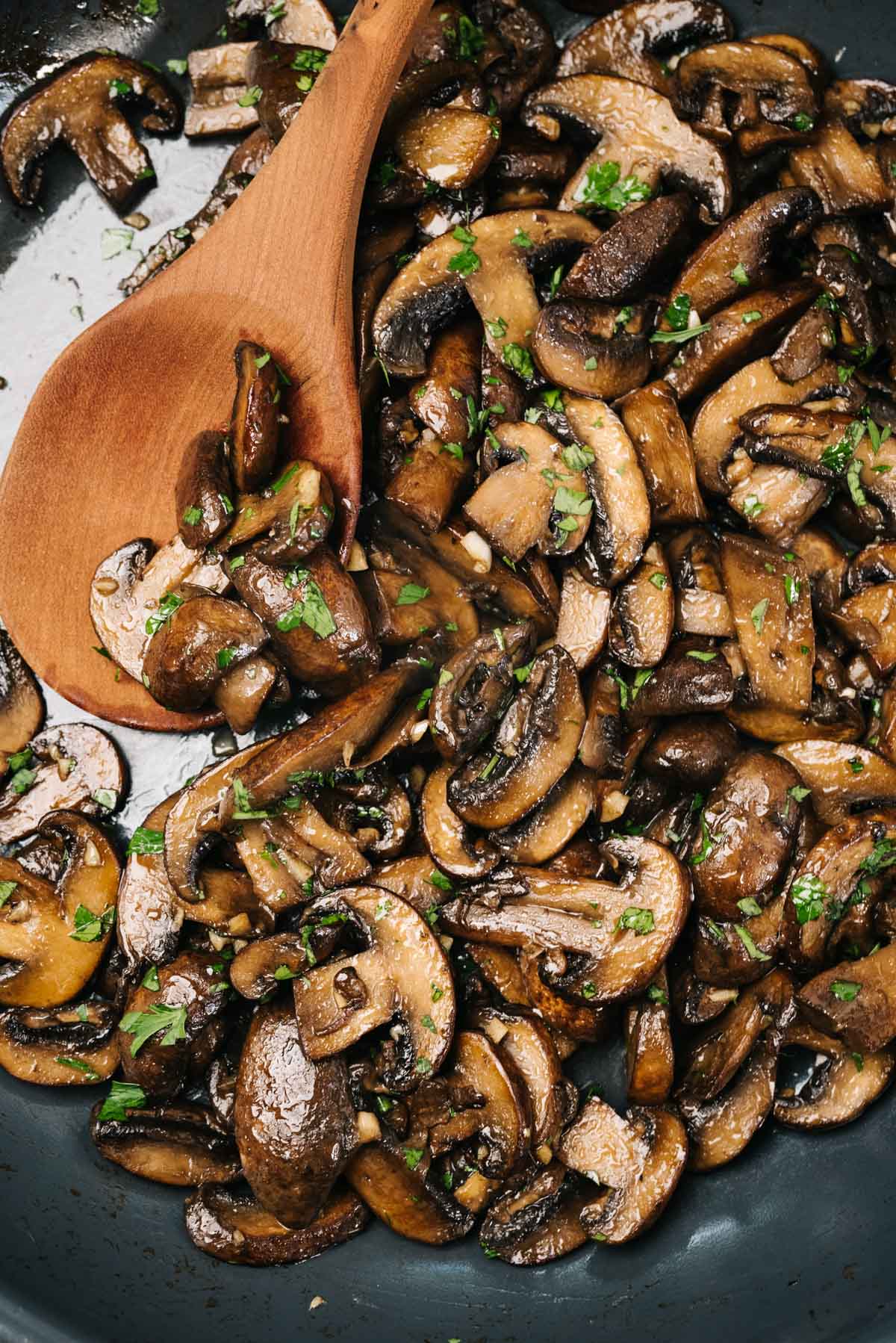 Detail of a wood spoon tucked into sautéed mushrooms garnished with fresh parsley in a skillet.