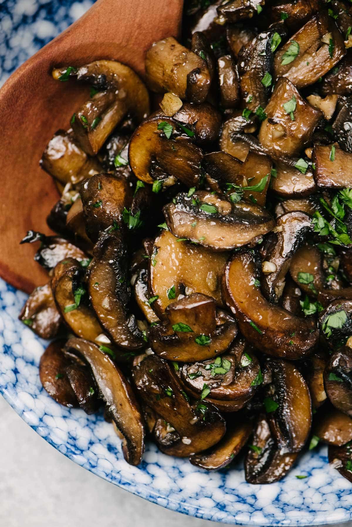 Detailed/close-up view of sauteed mushrooms in a blue speckled serving bowl with a wood spoon; the mushrooms are garnished with fresh chopped parsley.