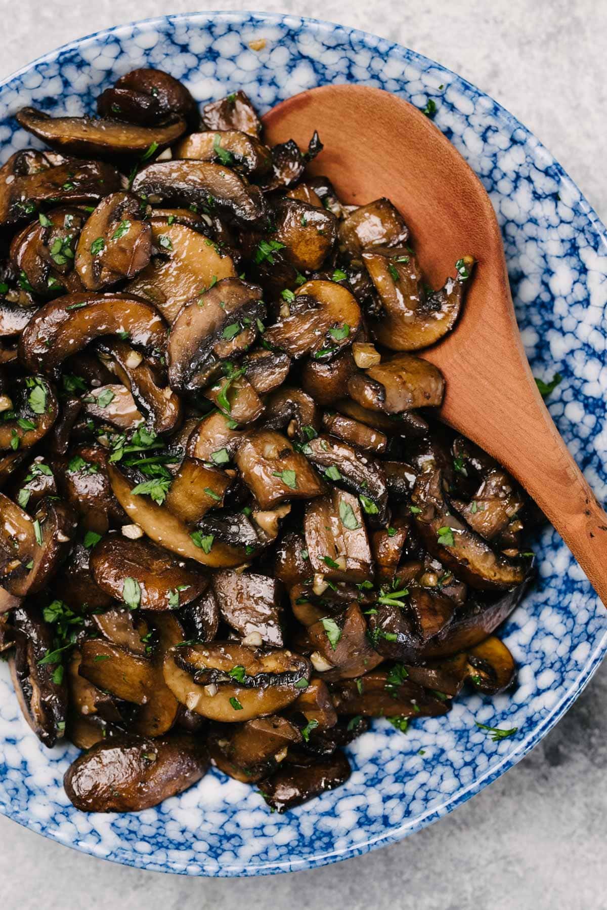 A wood spoon tucked into sauteed mushrooms in a blue speckled serving bowl on a cement background.