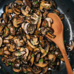 A wood spoon tucked into a skillet of golden brown sautéed mushrooms, garnished with fresh parsley.
