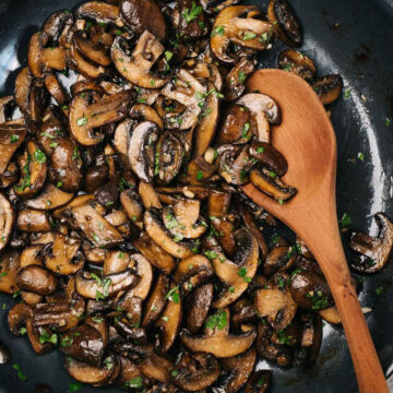 A wood spoon tucked into a skillet of golden brown sautéed mushrooms, garnished with fresh parsley.