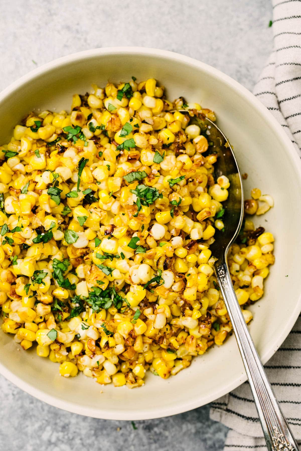 A spoon tucked into a bowl of sauteed skillet corn garnished with basil; striped linen napkin tucked to the side of the bowl.