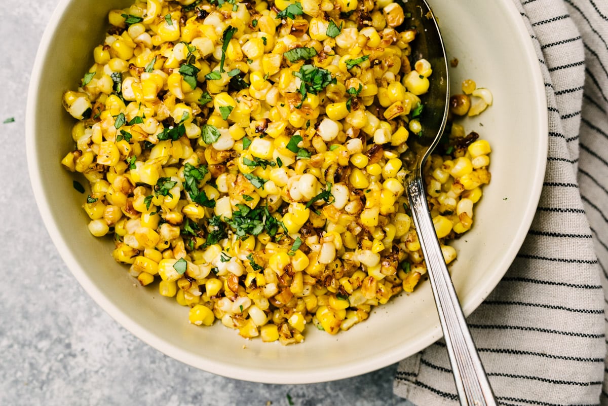 Skillet corn garnished with fresh basil in a low tan serving bowl; a spoon is tucked into the bowl, and a napkin is resting to the side.