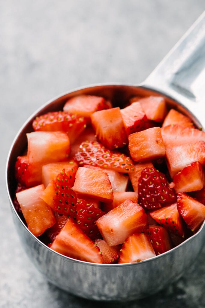 Side view, chopped strawberries in a one cup metal measuring cup on a concrete background.