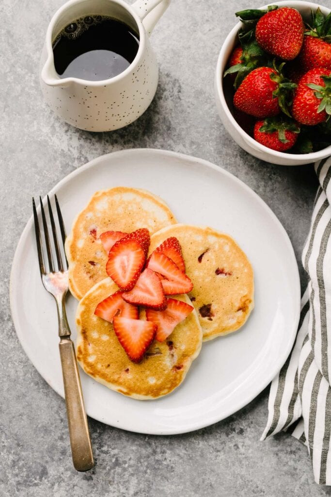 Three strawberry pancakes on a white plate with a silver fork, topped with sliced strawberries; linen napkin, pitcher of maple syrup, and bowl of fresh whole strawberries to the side.