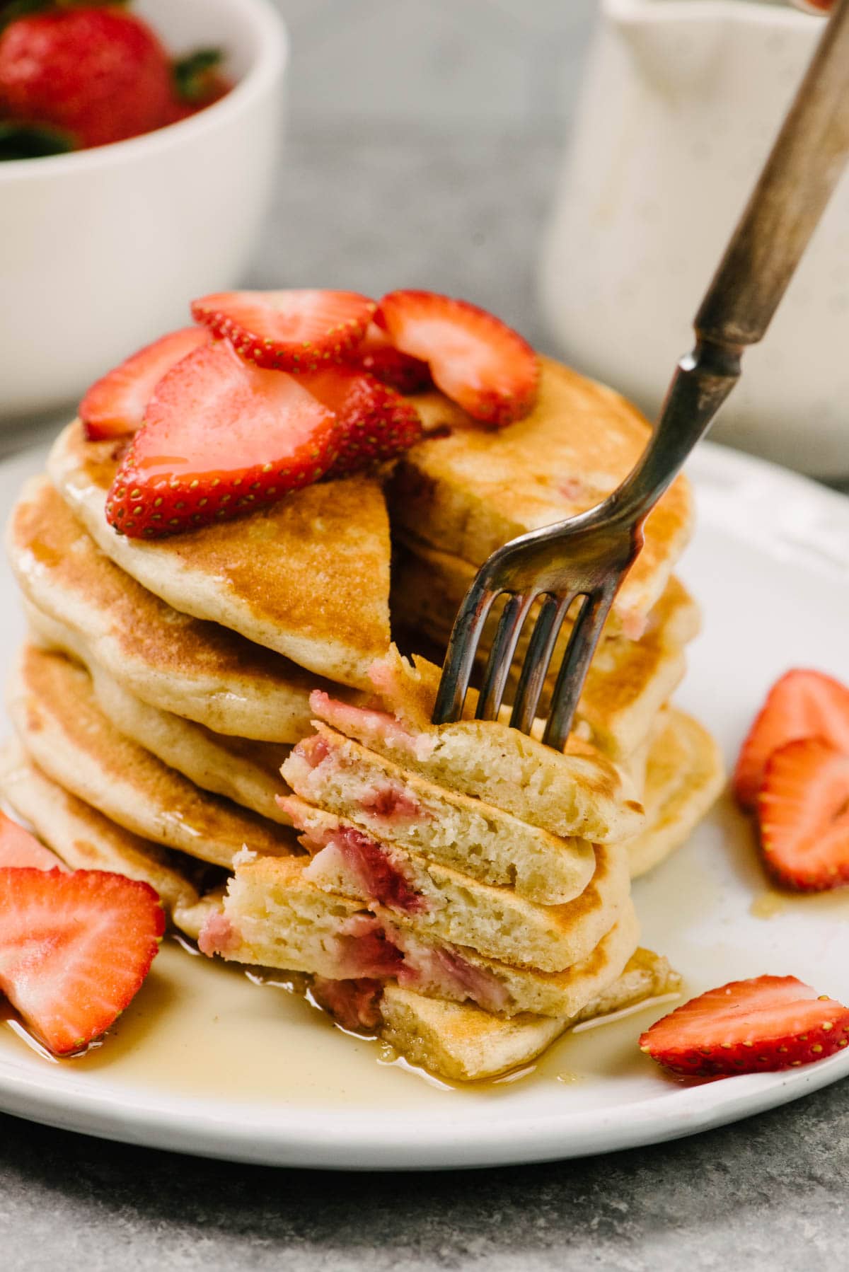 Side view, a fork removing a bite from a stack of strawberry pancakes on a white plate, with a pitch of maple syrup and bowl of fresh whole strawberries in the background.