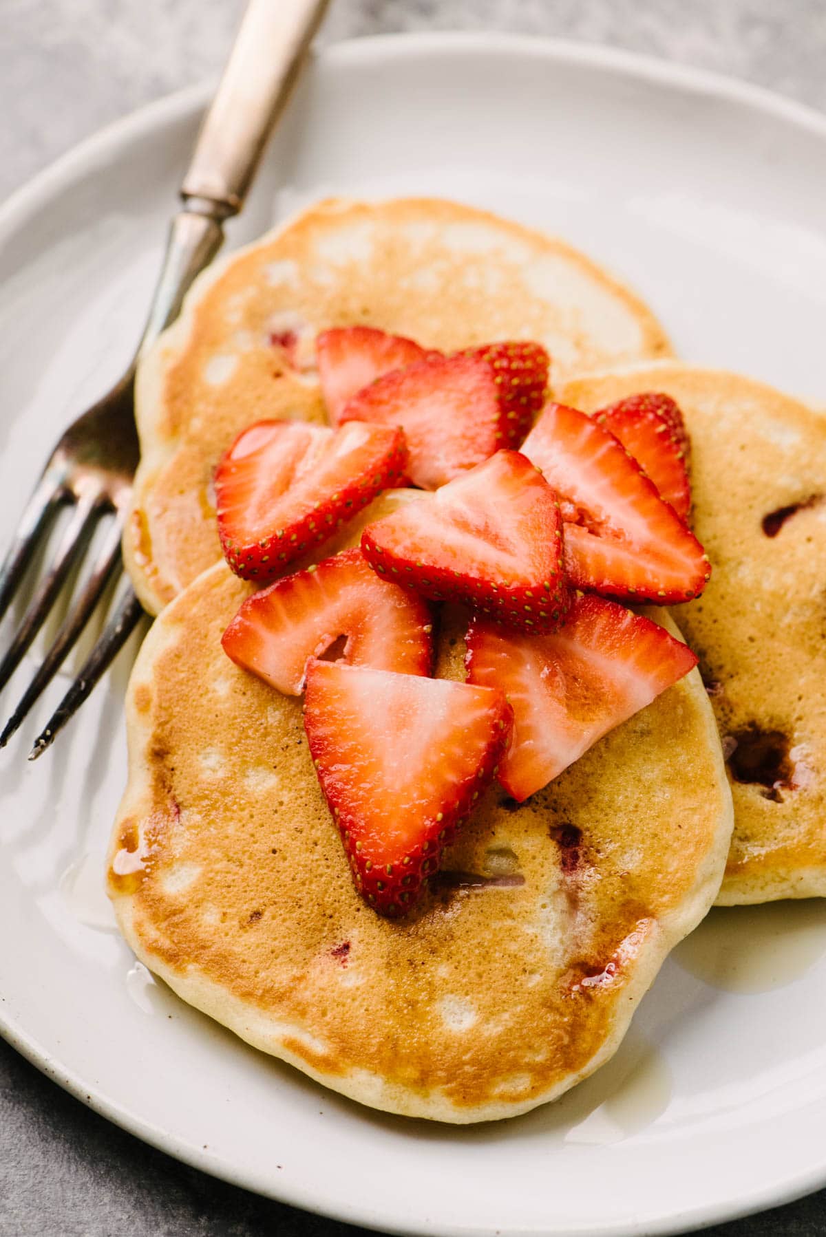 Side view, three strawberry pancakes on a white plate topped with sliced fresh strawberries; vintage silver fork to the side on the plate.