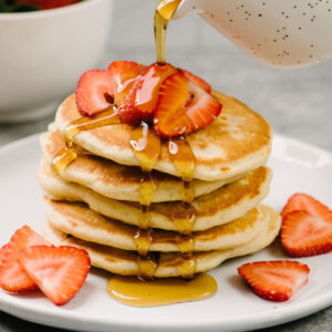Side view, pouring maple syrup over a stack of strawberry pancakes, topped with more fresh sliced strawberries; a bowl of fresh strawberries are in the background.