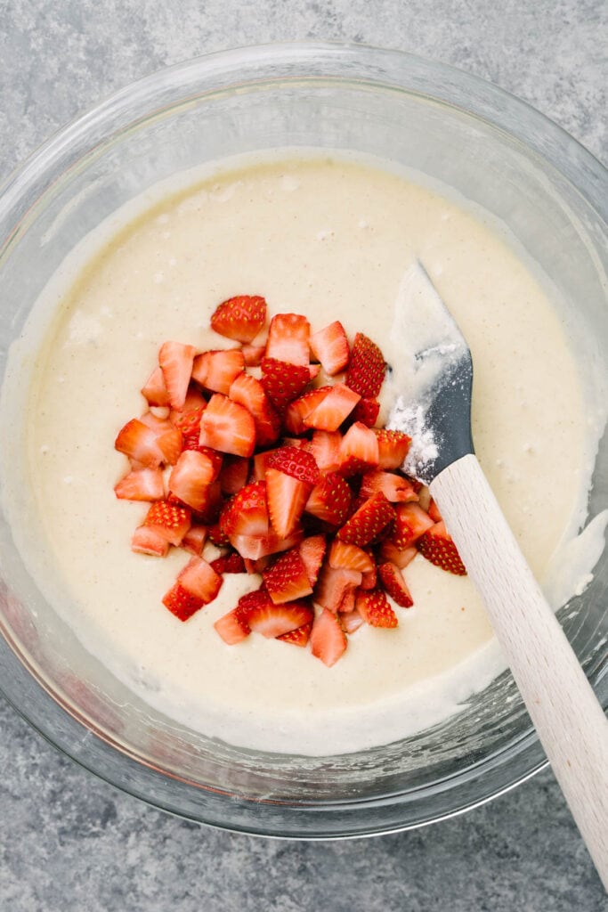 Adding fresh strawberries to pancake batter in a large glass mixing bowl with a rubber spatula.
