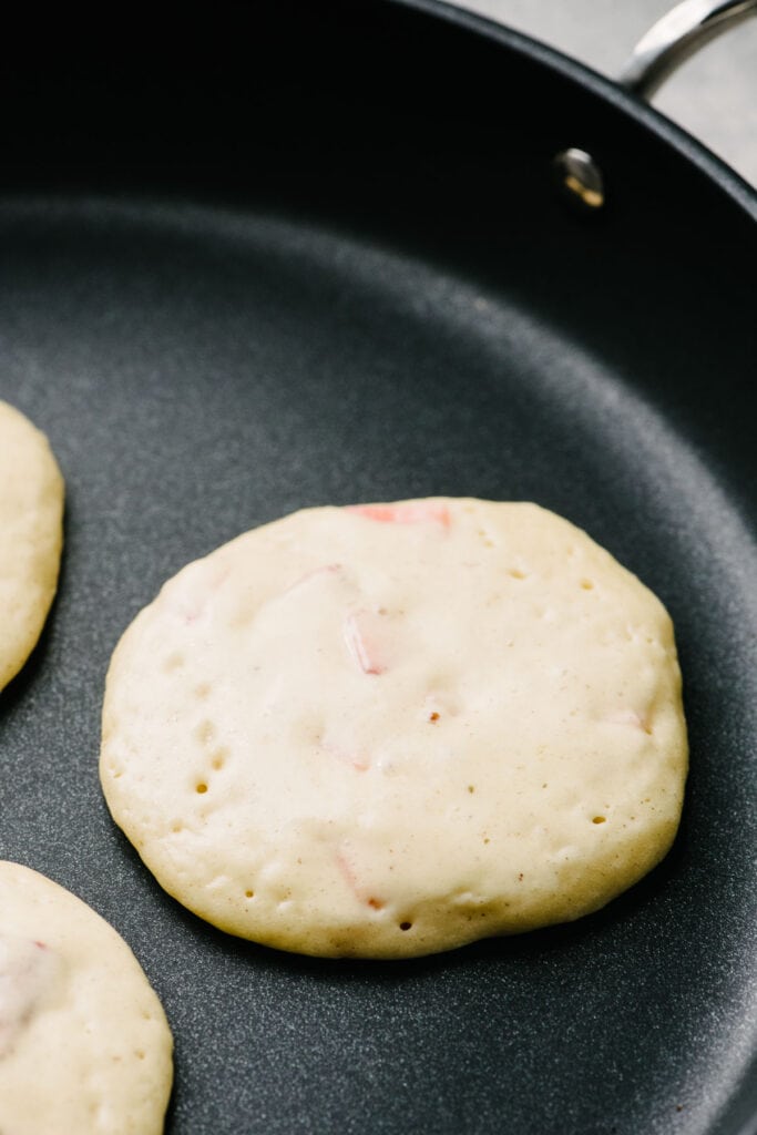 Side view, a strawberry pancake in a skillet before flipping showing the bubbles remaining on the surface and the firm edges.