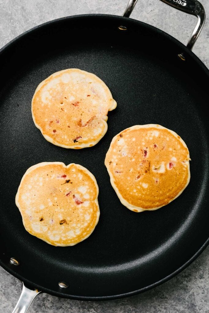 Three golden brown strawberry pancakes in a non-stick skillet.
