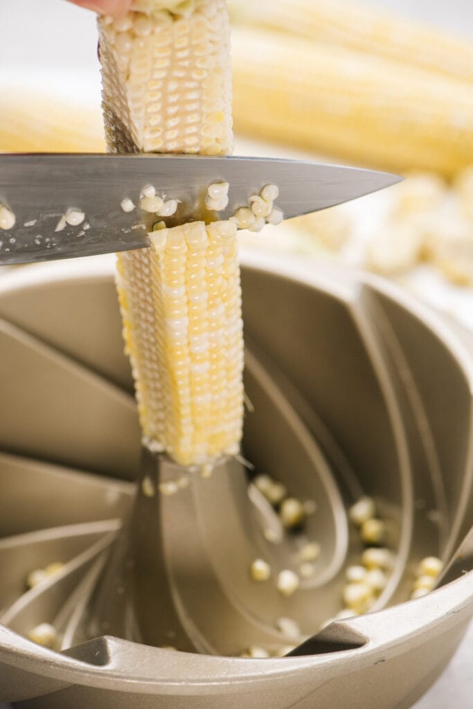 Side view, a hand holding a corn cob in the center of a bundt pan and slicing the kernels from the cob.