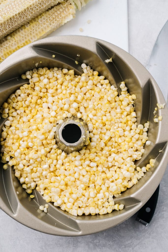 Corn kernels cut from the cobs in a bundt pan, surrounded by trimmed cobs and a chef's knife.