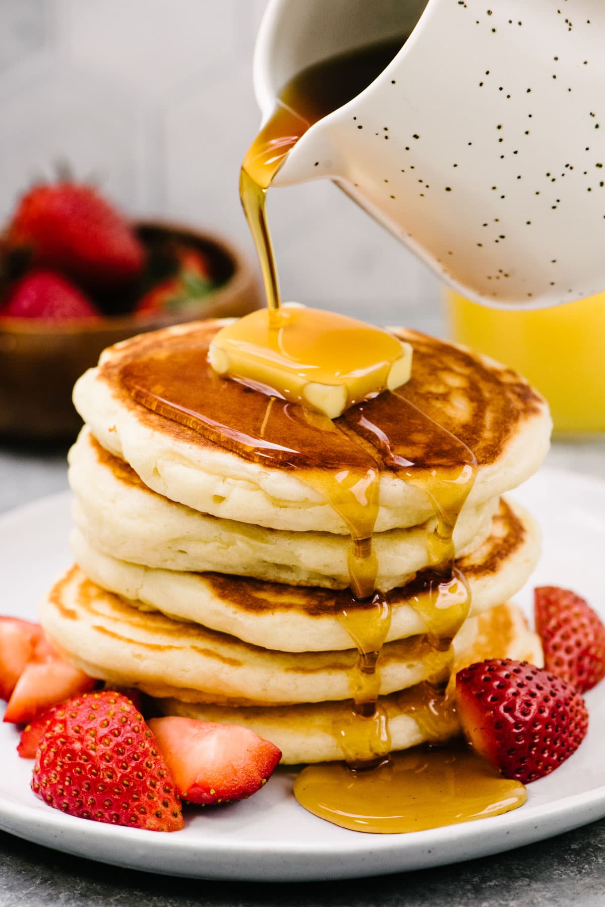 Side view, pouring maple syrup from a small speckled pitcher over a stack of fluffy homemade pancakes topped with butter and plated with fresh strawberries.
