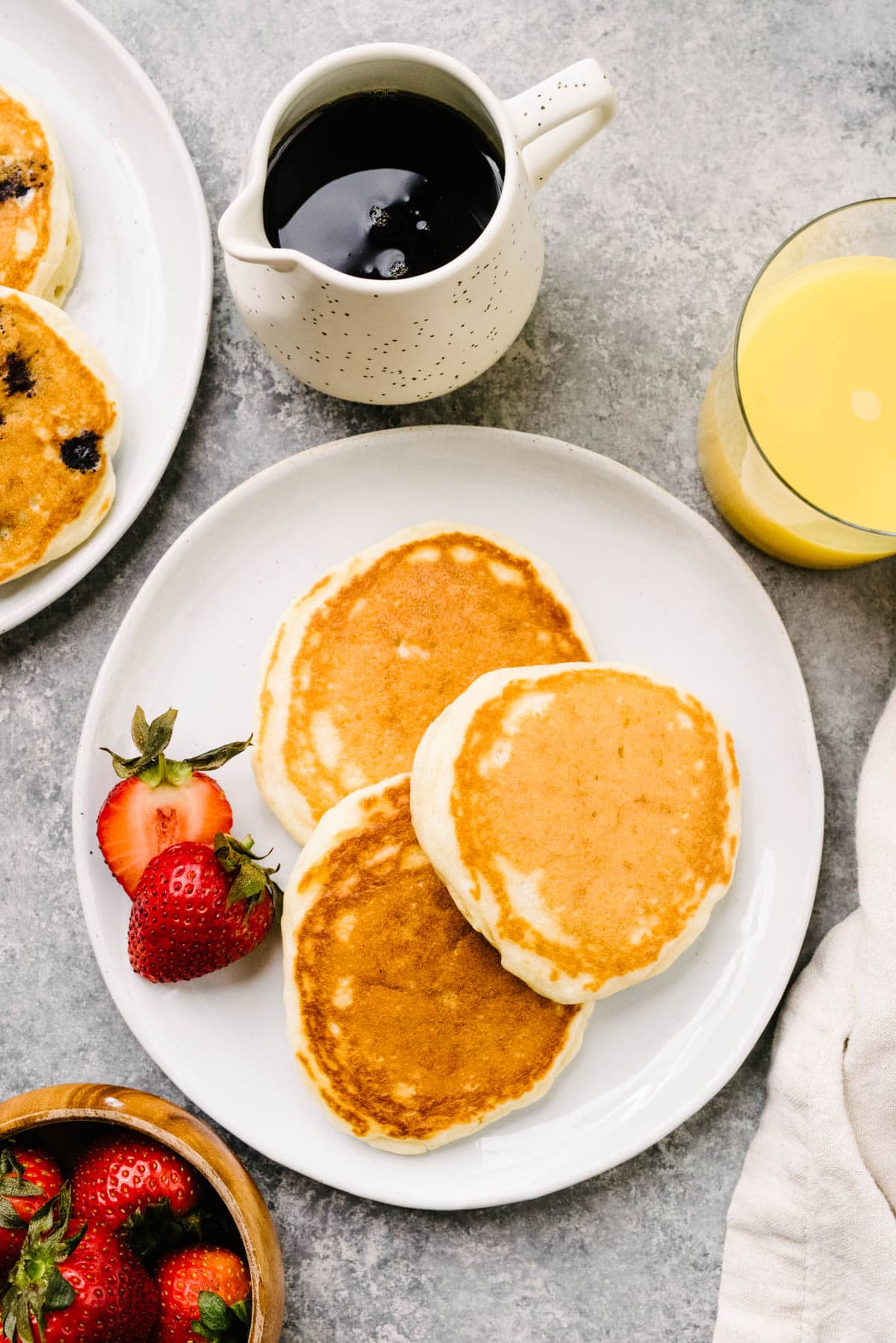 Three homemade pancakes on a white plate with strawberries; a small pitcher of maple syrup, glass of orange juice, bowl of strawberries, and cream linen napkin surround the plate.