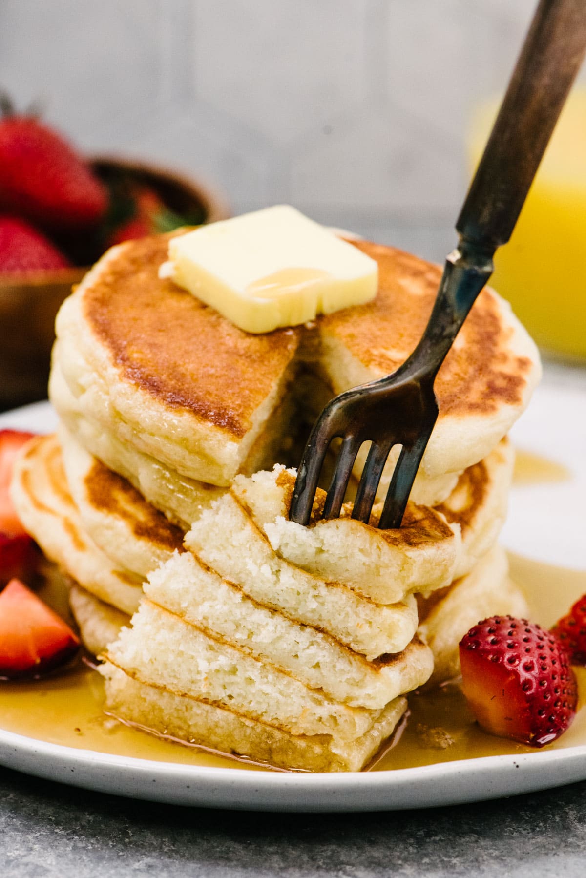 Side view, a fork removing a bite from a stack of pancakes topped with butter and drizzled with maple syrup.