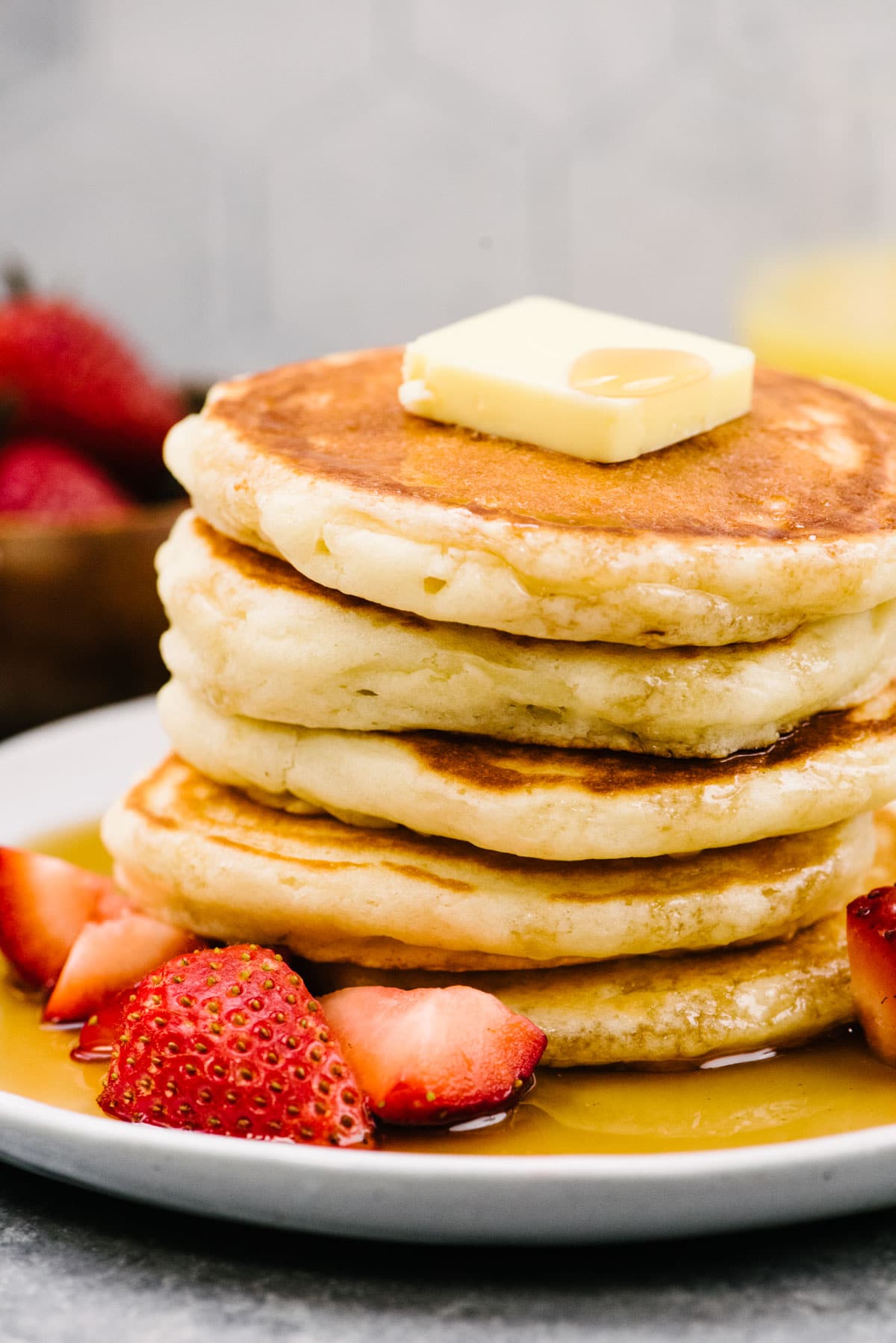 Side view, a stack of homemade pancakes on a white plate topped with butter, drizzled with maple syrup, and garnished with fresh strawberries.