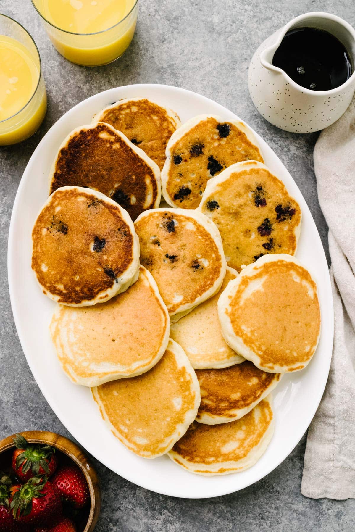 A platter of plain homemade pancakes, blueberry pancakes, and chocolate chip pancakes on a marble background; two glasses of orange juice, a bowl of strawberries, a small pitcher of maple syrup, and cream linen napkin surround the platter.