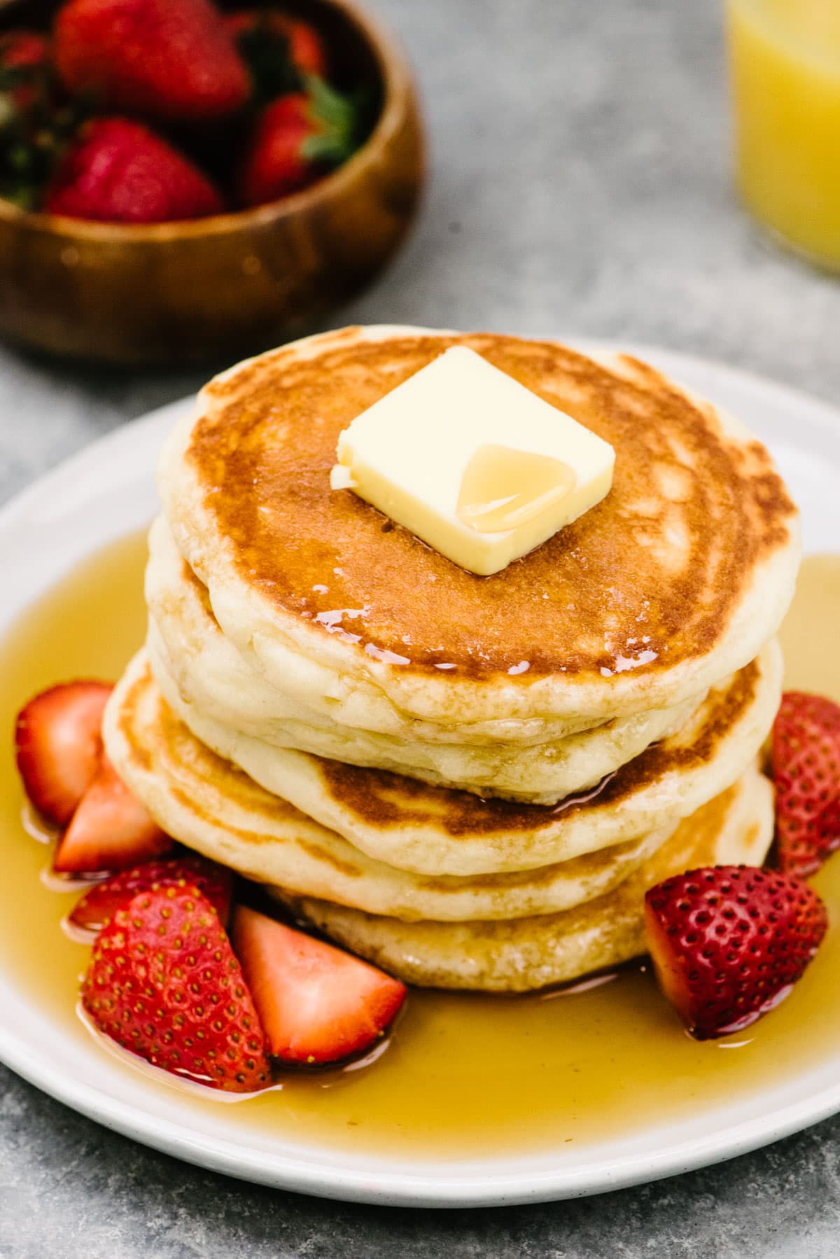 Side view, a stack of homemade fluffy pancakes on a white plate topped with butter and drizzled with maple syrup. The plate has sliced strawberries to the side. A glass of orange juice and bowl of fresh strawberries in the background.