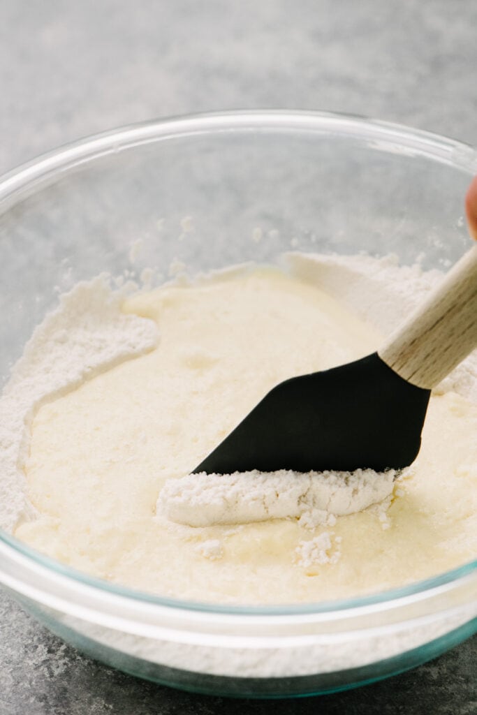 Side view, gently folding wet ingredients into dry ingredients in a large glass mixing bowl to make homemade pancake batter.