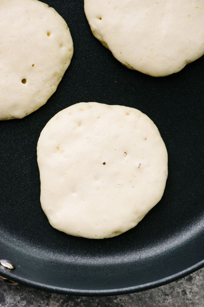 Three pancakes cooking in a non-stick skillet showing the bubbles in the batter before flipping.